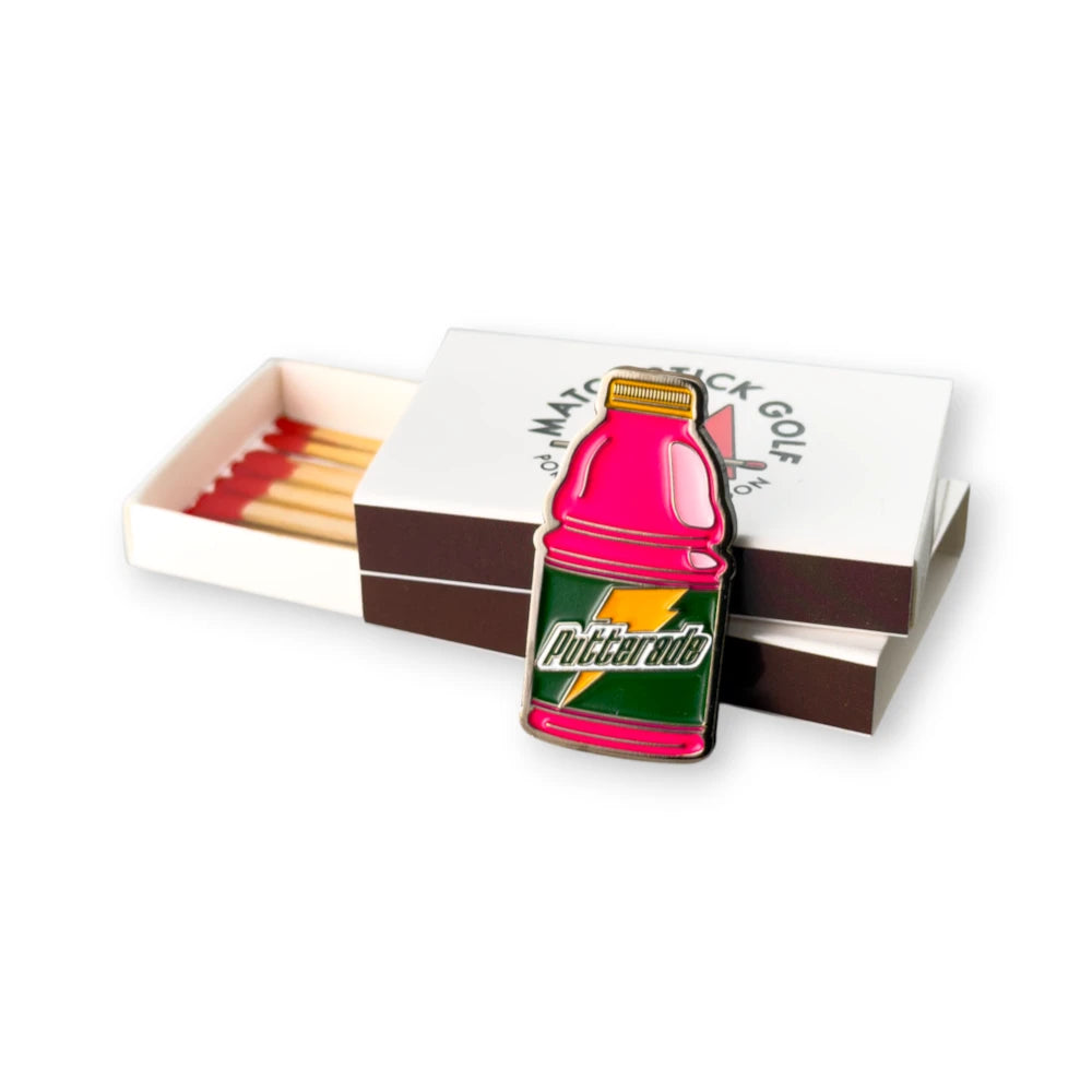 pink putterade golf ball marker leaned up against stacked matchbox packaging