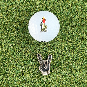 black and gold devil horns golf ball marker  on grass next to white golf ball