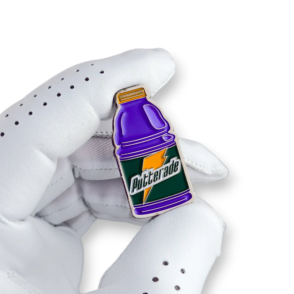 A small enamel golf ballmarker shaped like a Putterade gatorade sports drink bottle, held between a white perforated golf glove and fingers against a clean white background.